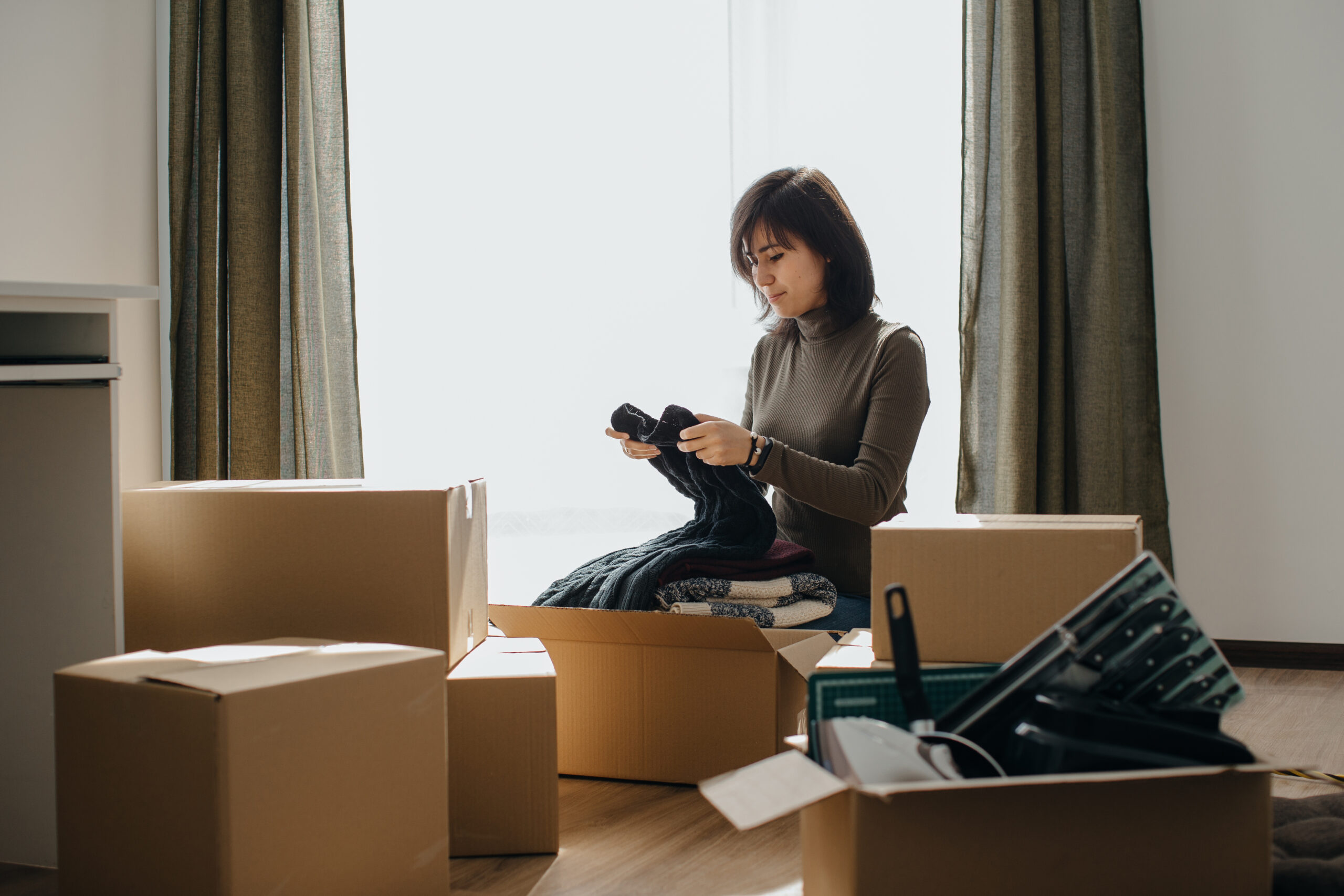 Woman unpacking clothes from a cardboard box in her new apartment, surrounded by moving boxes and embracing the excitement of relocation